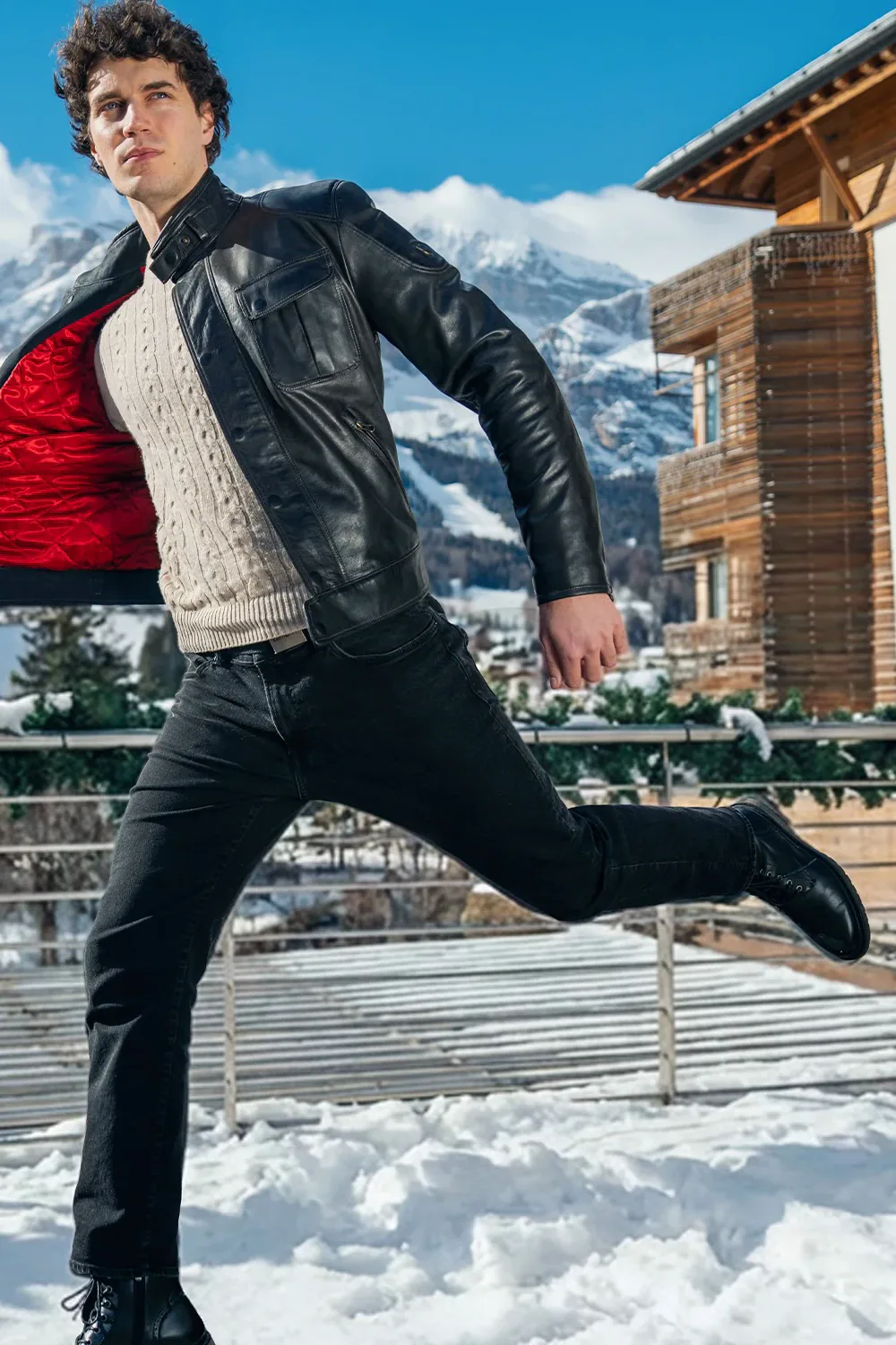 Francesco Scarparo in a leather jacket, mid-air over the snow, with the Dolomites behind him